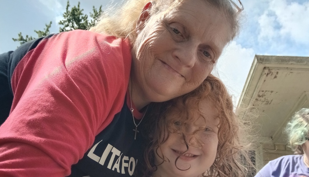 A smiling older woman leaning close to a young child with curly red hair outdoors.