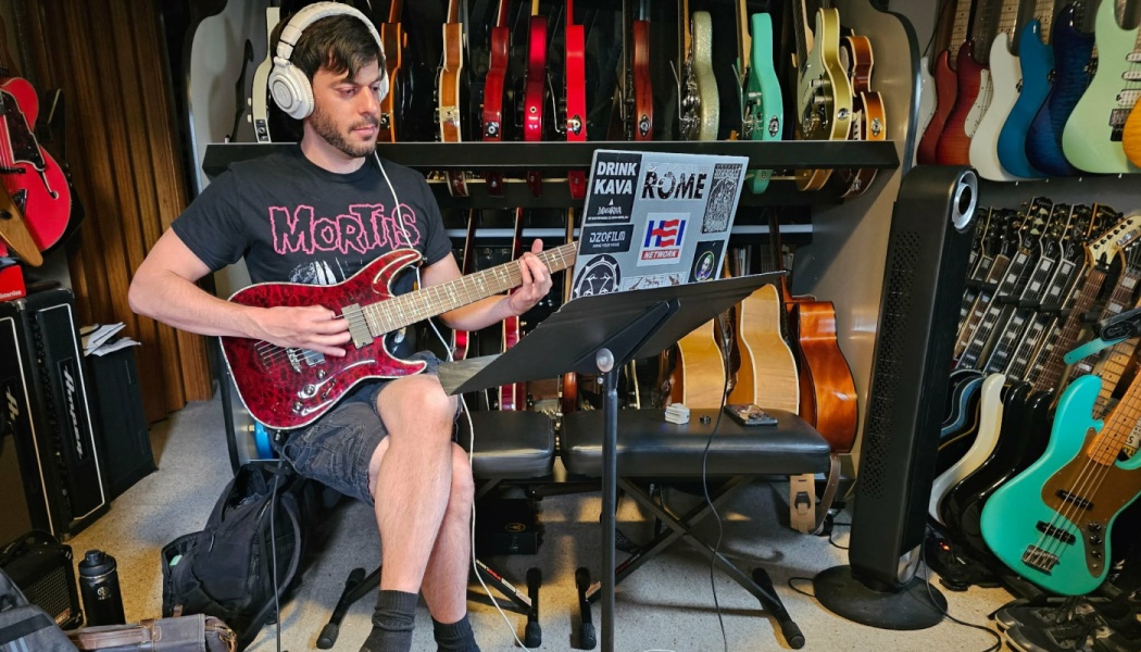 A man wearing headphones plays a red electric guitar in front of a laptop in a studio filled with guitars.