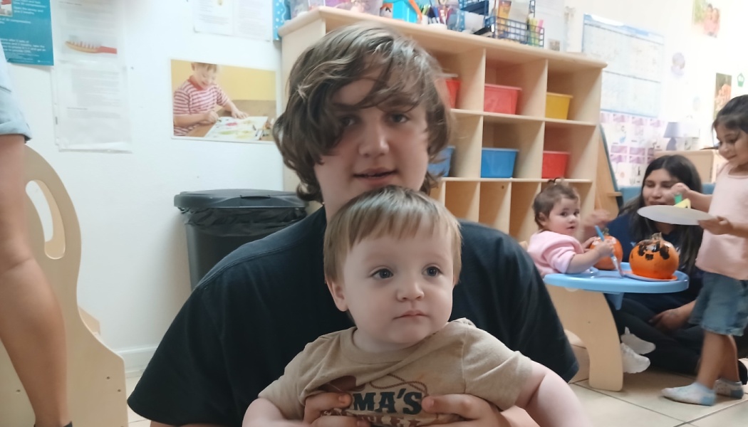 A teenage boy holding a young toddler in a daycare classroom with other children playing behind them.