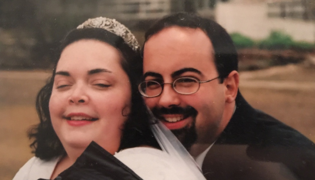 A smiling bride wearing a tiara poses closely with her smiling groom who wears glasses and a goatee.