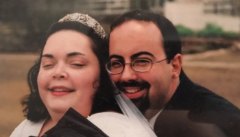 A smiling bride wearing a tiara poses closely with her smiling groom who wears glasses and a goatee.