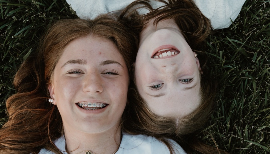 Two cheerful young girls lying head-to-head on green grass, smiling at the camera.