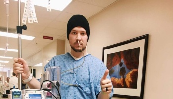 Man in a hospital gown, nasal tube, and beanie giving a thumbs-up while holding an IV pole in a hospital hallway.