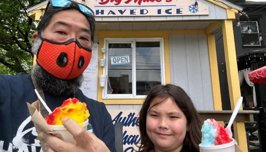 A man wearing an orange mask and a child holding colorful shaved ice outside a shaved ice stand.