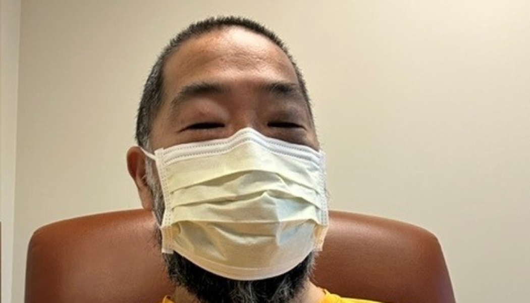 Close-up of a man with short hair and a beard wearing a white medical mask while sitting in a brown chair.