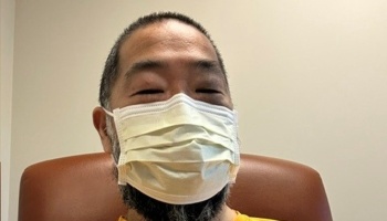 Close-up of a man with short hair and a beard wearing a white medical mask while sitting in a brown chair.