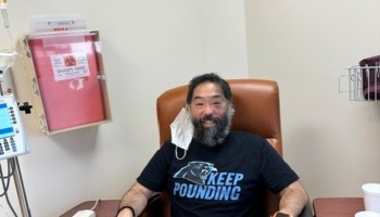 A smiling bearded man wearing a "Keep Pounding" t-shirt sits in a medical treatment chair beside IV equipment.