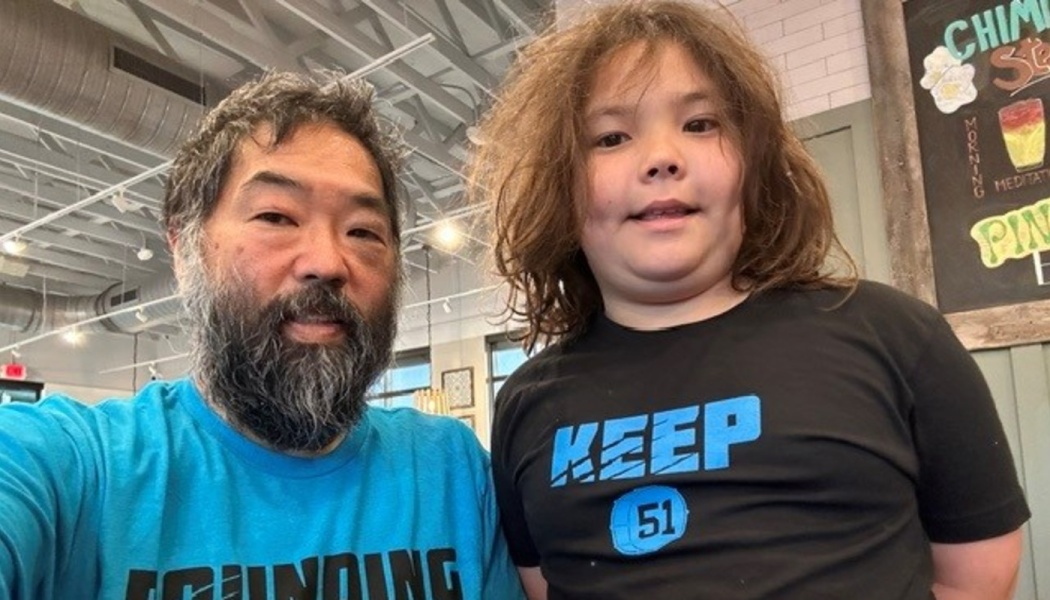 A bearded man in a bright blue t-shirt posing close next to a smiling child with long brown hair indoors.