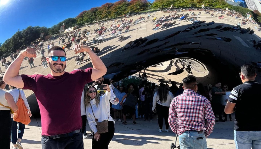 A smiling man in sunglasses flexes his biceps in front of Chicago's reflective Cloud Gate sculpture.