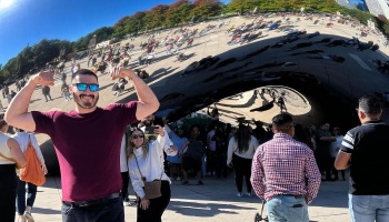 A smiling man in sunglasses flexes his biceps in front of Chicago's reflective Cloud Gate sculpture.
