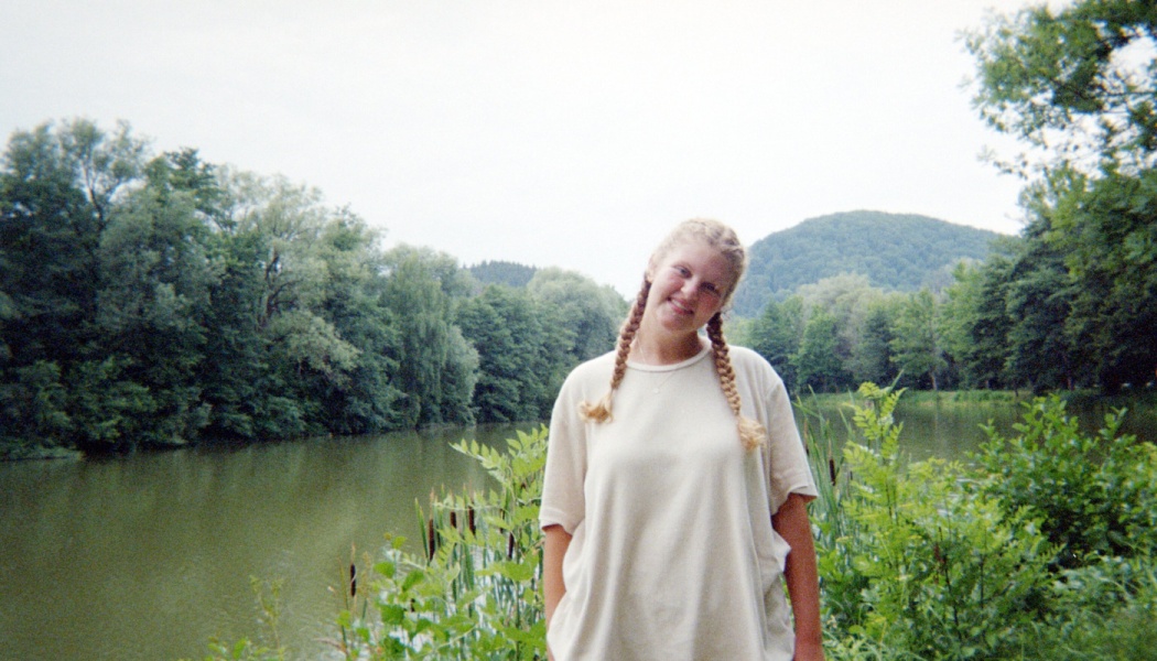 A smiling young woman with braided hair stands beside a calm lake surrounded by lush green trees and hills.