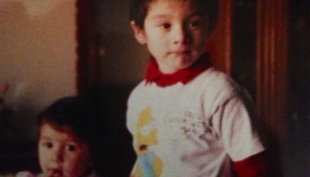 A young boy wearing a white graphic shirt with a red collar stands beside a toddler looking toward the camera.
