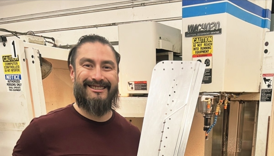 A smiling bearded man holds a large machined metal part next to a vertical CNC machine in a workshop.