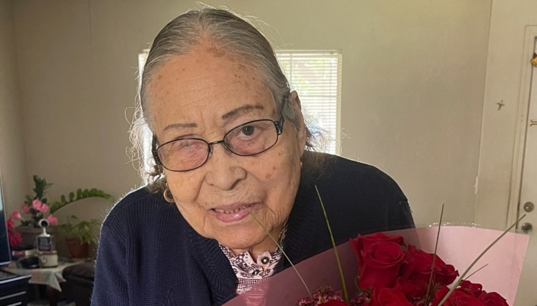An elderly woman wearing glasses smiles while holding a bouquet of red roses.