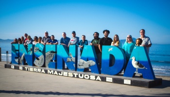 A group of people poses behind large blue letters spelling ENSENADA along a sunny oceanfront promenade.