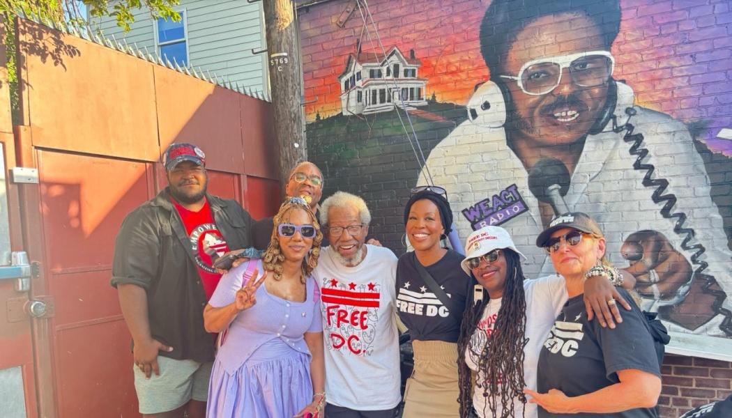 Seven smiling people in Free DC apparel pose together outdoors in front of a WE ACT Radio brick wall mural.