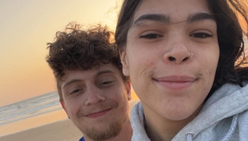 Close-up selfie of two smiling young people on a beach at sunset.