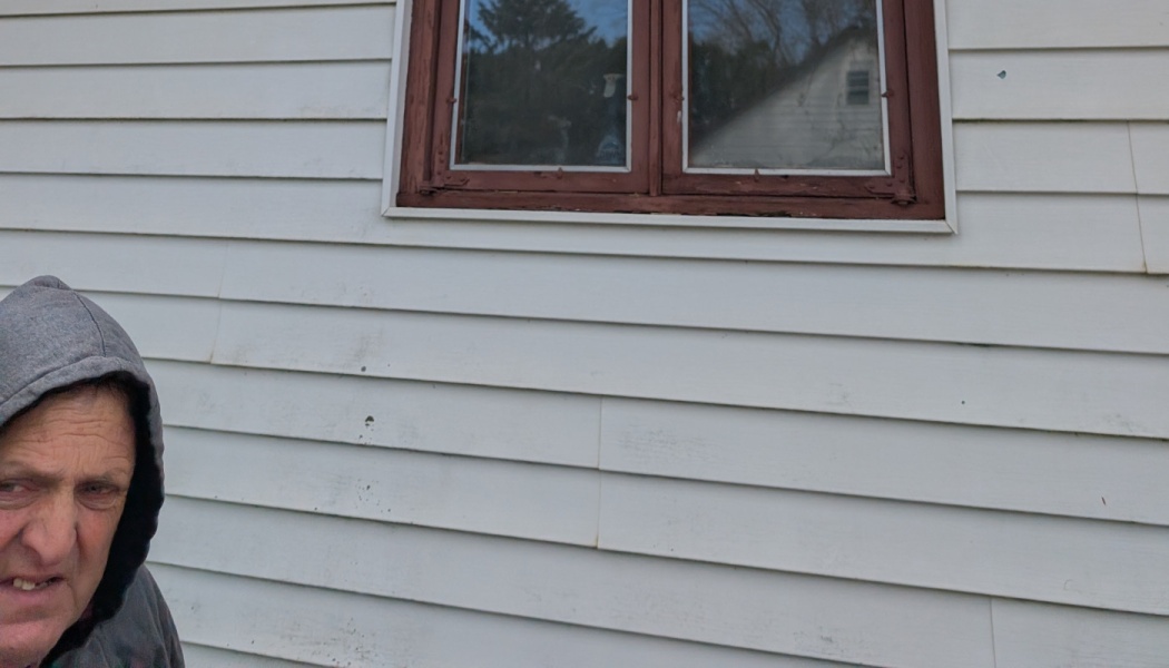 A man in a grey hoodie standing outside beside white house siding under a brown wooden window.