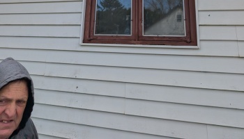 A man in a grey hoodie standing outside beside white house siding under a brown wooden window.
