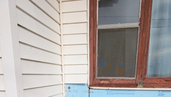 Weathered wooden window frame with peeling paint next to white siding and exposed blue insulation foam.