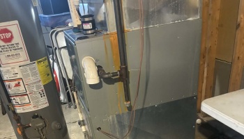 An old HVAC furnace with visible rust stains standing next to a water heater in an unfinished basement.