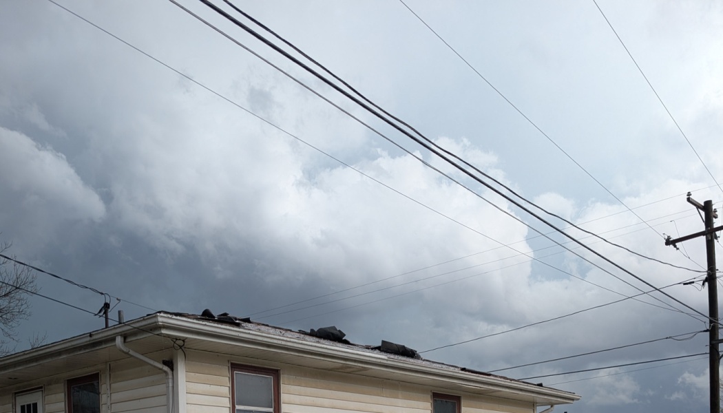 Damaged roof shingles on a house under dark storm clouds and crisscrossing power lines.