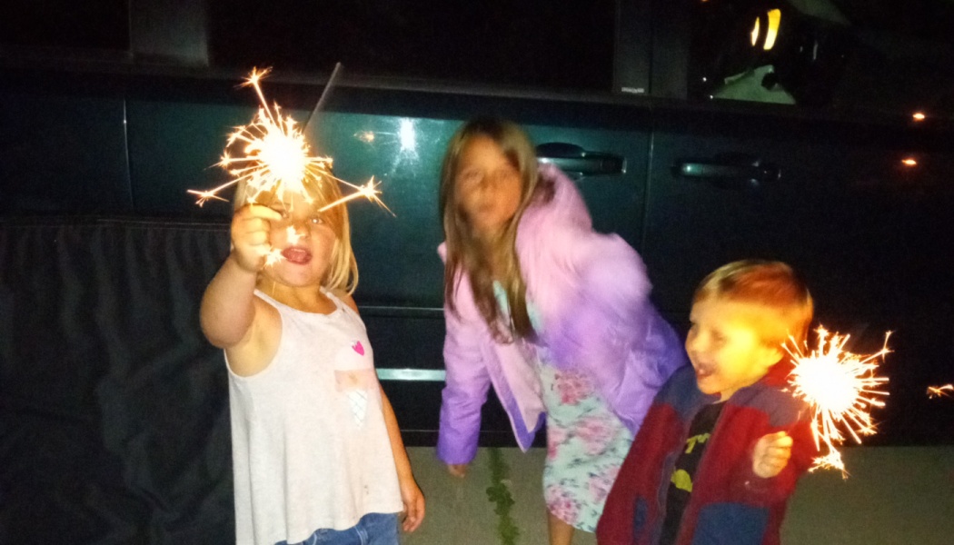 Three young children holding glowing sparklers and smiling outdoors at night.