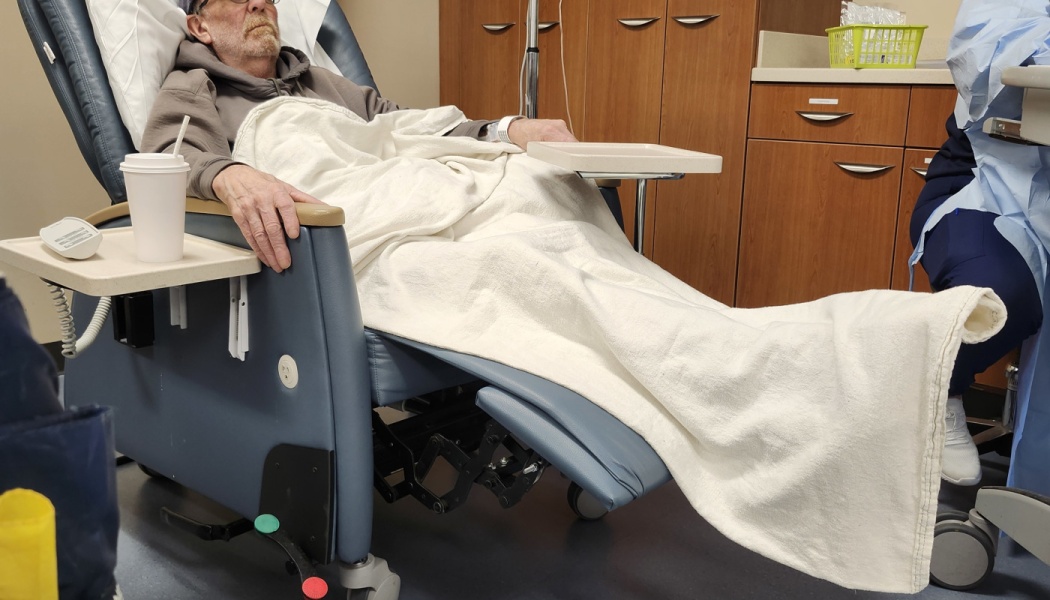 An elderly man rests in a reclining hospital chair covered with a white blanket during treatment.