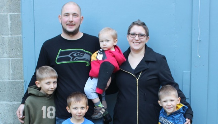 A smiling family of six, including two parents and four young boys, posing together against a light blue backdrop.
