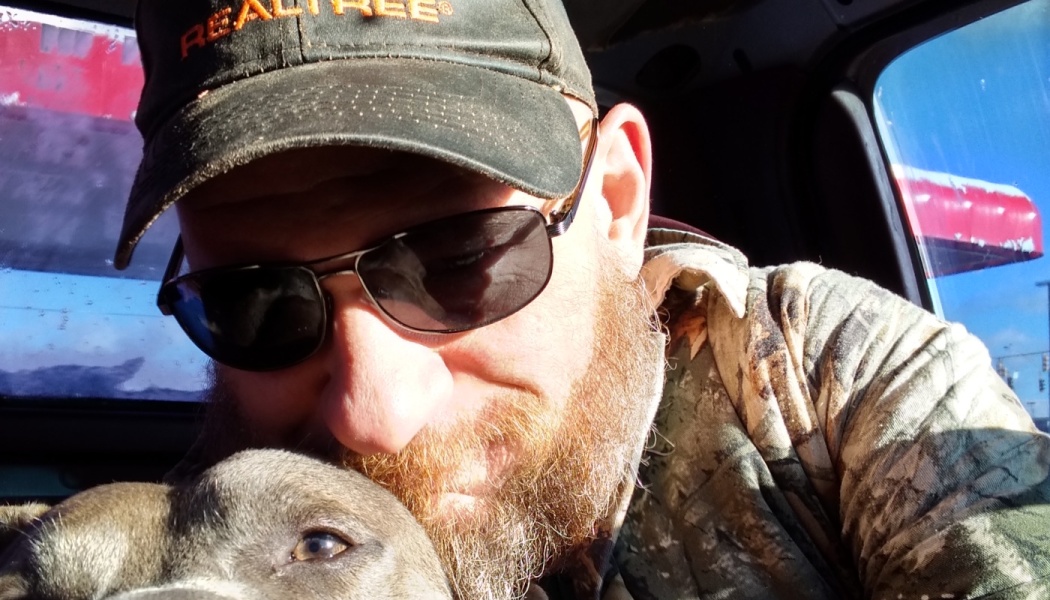 A bearded man in sunglasses and a camo hat rests his head affectionately against his dog inside a vehicle.