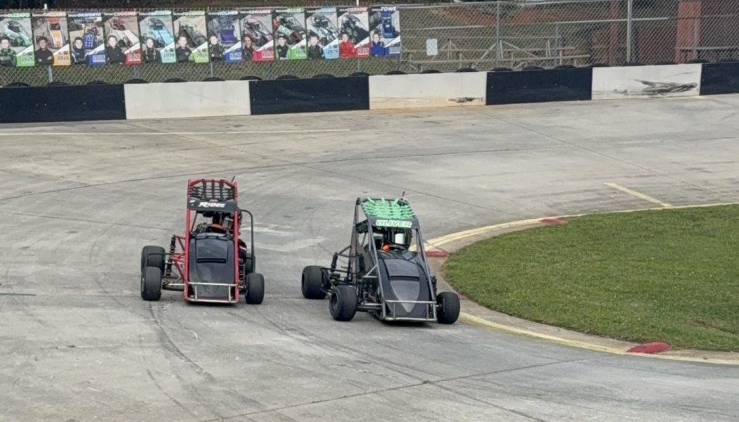 Two youth sprint cars race side-by-side around a curve on a paved racetrack.