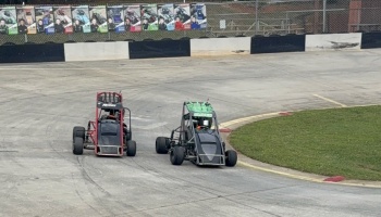 Two youth sprint cars race side-by-side around a curve on a paved racetrack.
