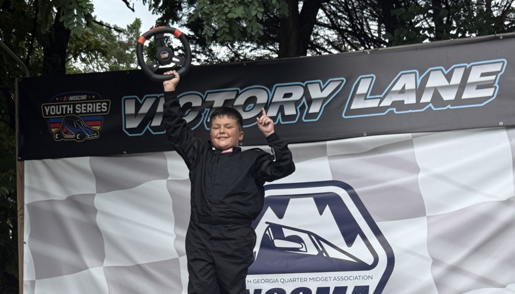 A smiling young boy in a black racing suit holds up a steering wheel in front of a Victory Lane banner.