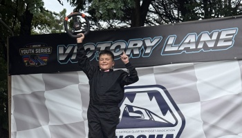 A smiling young boy in a black racing suit holds up a steering wheel in front of a Victory Lane banner.