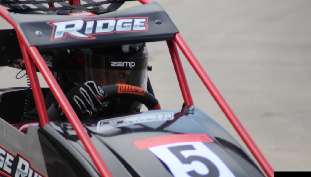 A helmeted race car driver behind the wheel of a black number 5 race car with a red roll cage.