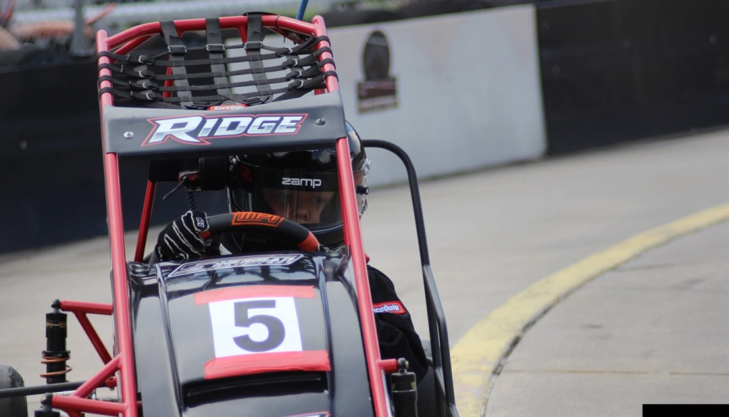 A young driver wearing a black helmet sits behind the wheel of a red and black number 5 mini race car on a track.