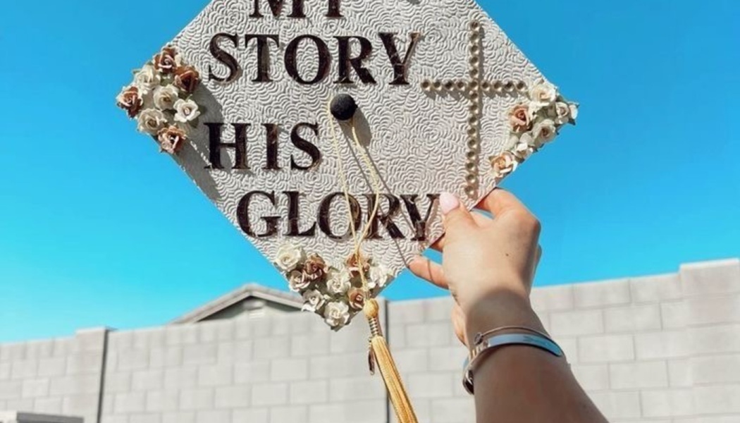Hand holding a decorated graduation cap reading "MY STORY HIS GLORY" with flowers and a cross against a blue sky.