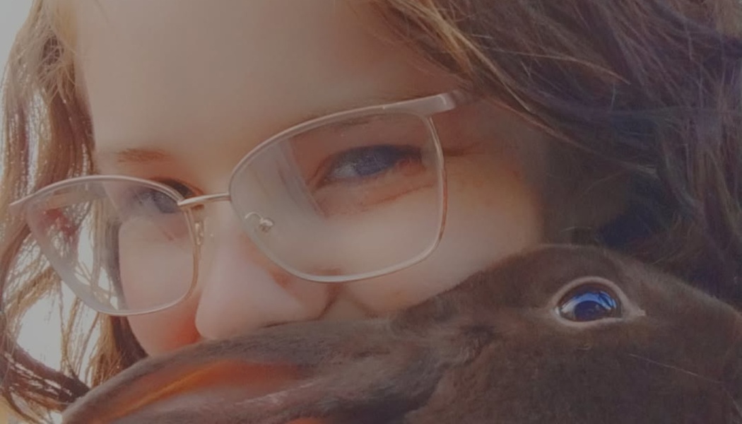 Close-up of a young woman wearing glasses holding a dark brown rabbit close to her face.