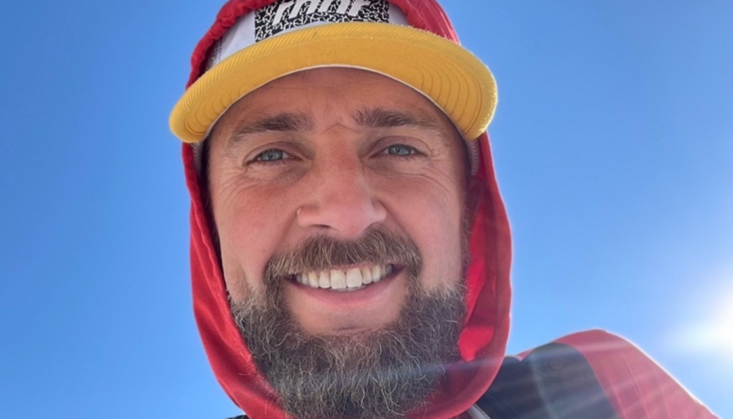 A smiling bearded man wearing a red hoodie and yellow-brimmed cap against a clear blue sky.