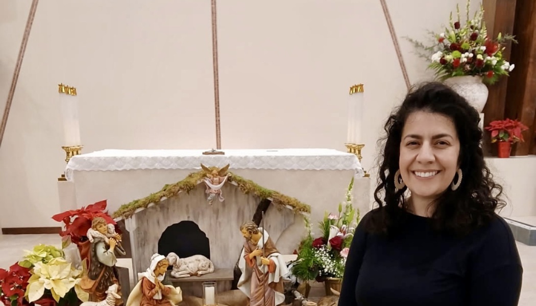 A smiling woman with dark curly hair stands in front of a church altar with a nativity scene.