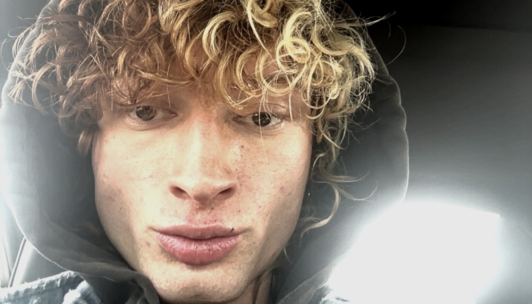 Close-up selfie of a young man with curly blonde hair wearing a dark hood pulled over his head.