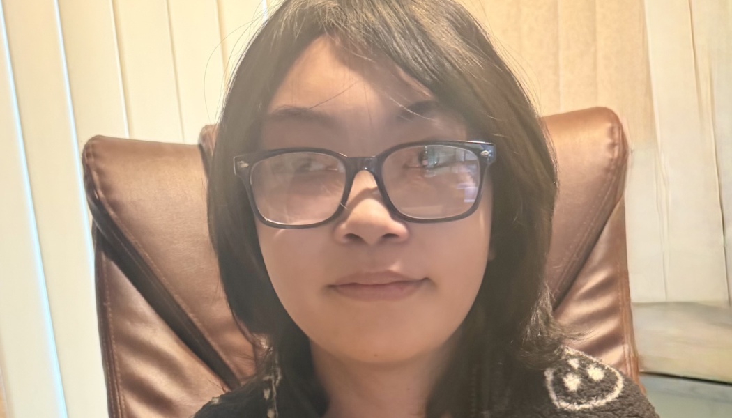 A young person with dark shoulder-length hair and black glasses sitting in a brown leather chair.