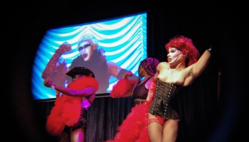Performers in red boas and dramatic costumes dance on stage in front of a glowing projection screen.