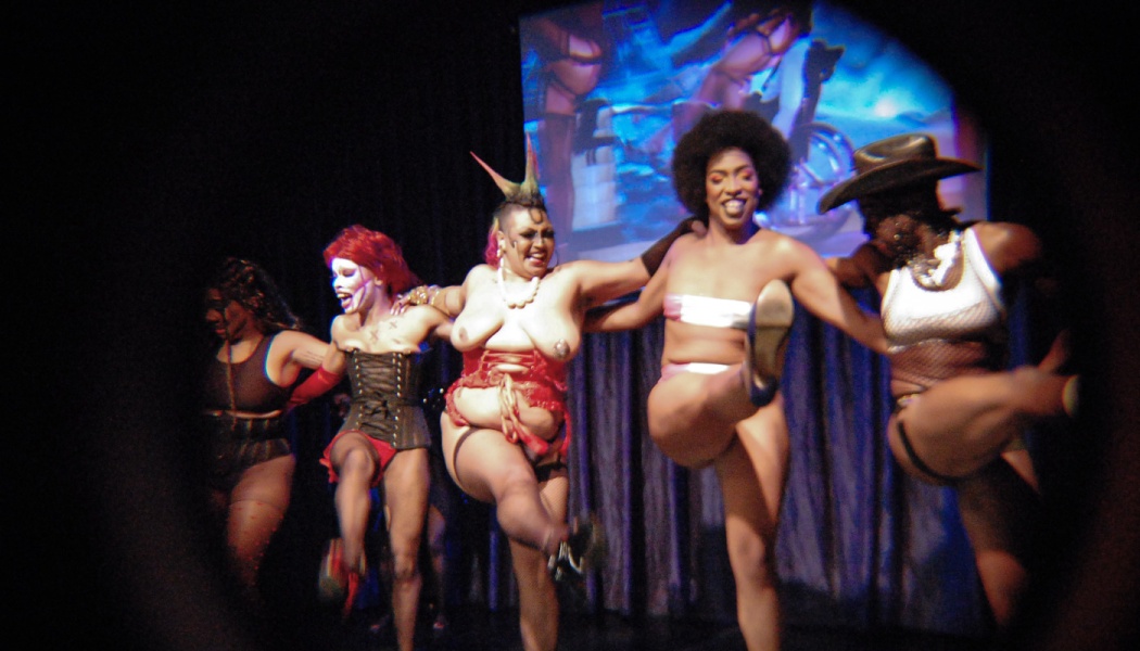 Diverse burlesque performers link arms and kick their legs high in unison during a stage show.