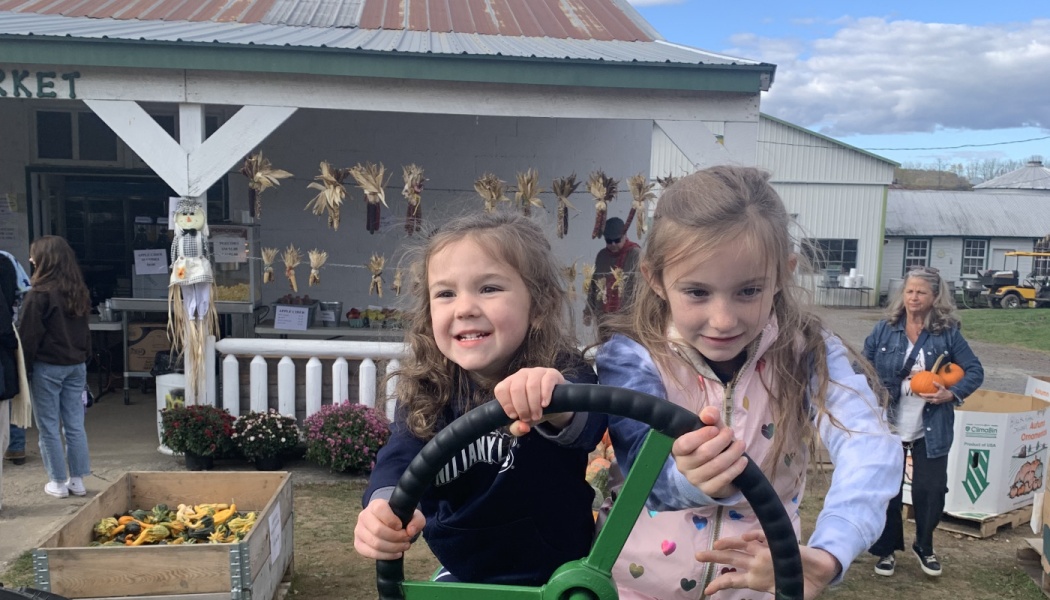 Two smiling young girls hold a large steering wheel at an outdoor autumn farm market.
