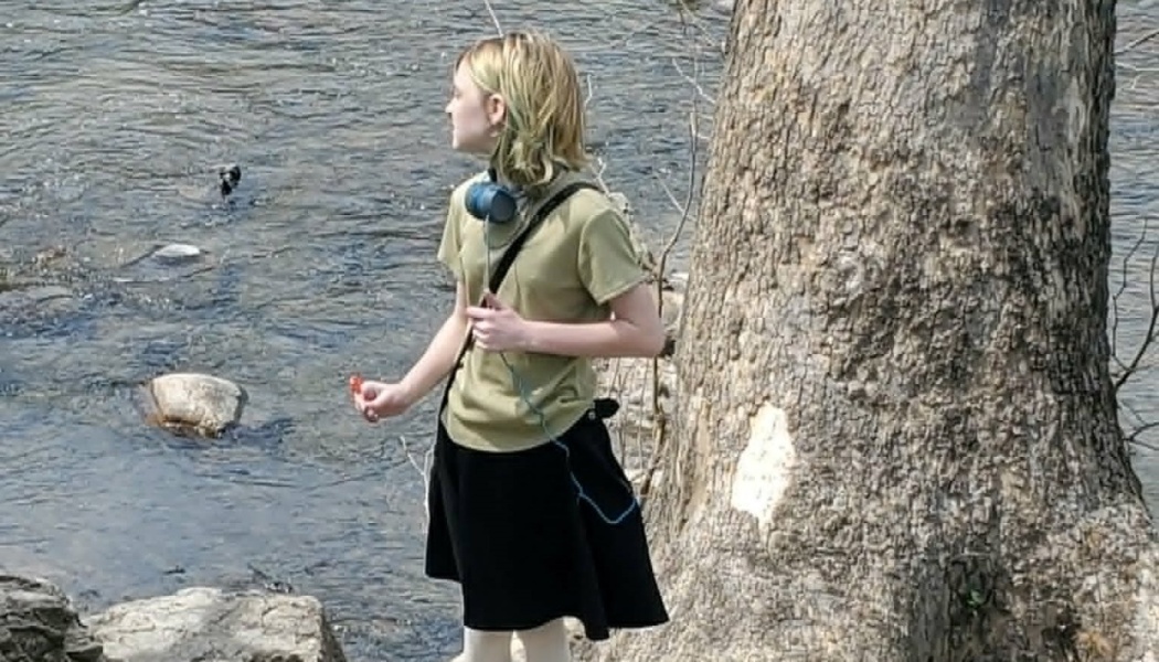 Young person with headphones standing by a river next to a large tree trunk.
