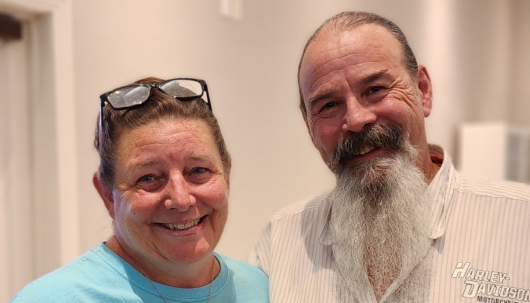 A smiling couple standing together: a woman with glasses on her head and a man with a long white beard.