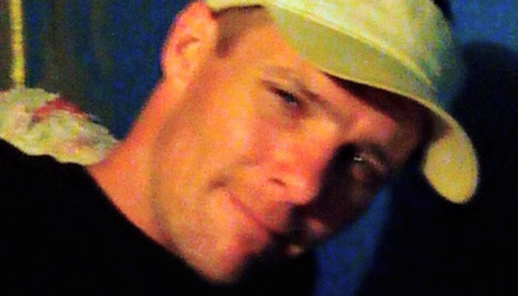 Close-up of a smiling man wearing a light-colored baseball cap and dark shirt.