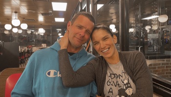 A smiling man and woman sit close together in a diner booth, her arm lovingly wrapped around his neck.