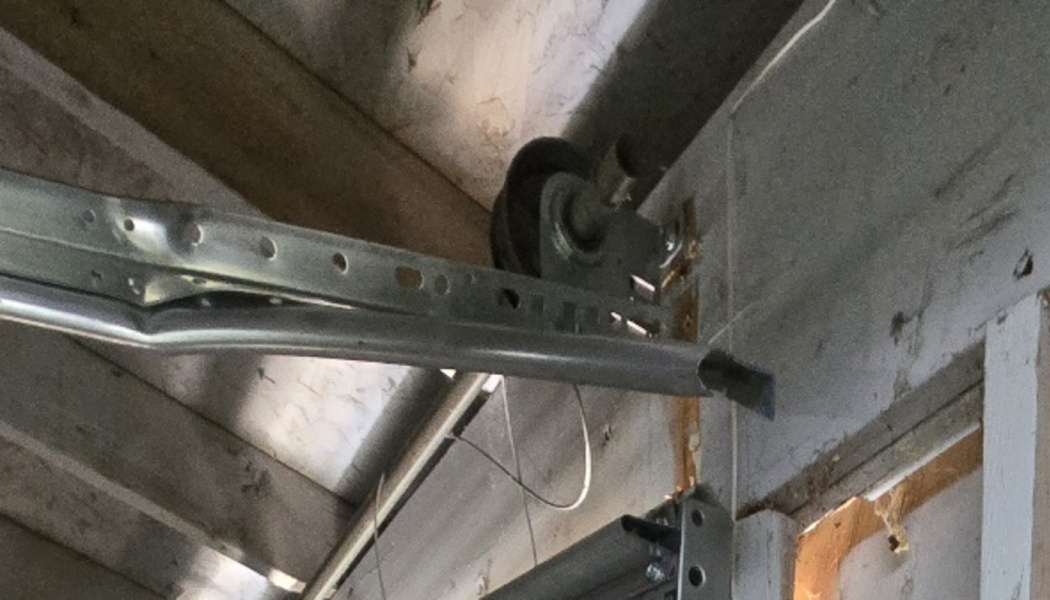 Severely bent and damaged metal garage door track and pulley mounted to wooden ceiling rafters.